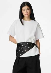 Wit oversized T-shirt met korte mouwen, gecombineerd met een zwarte ceintuurachtige wikkel met stippen die de taille accentueert. Zwarte broek maakt de look compleet.