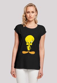 Svart t-skjorte med en gul tegneseriekanarifugl med blå øyne, stående med hendene på hoftene, i en rundhalsedesign.