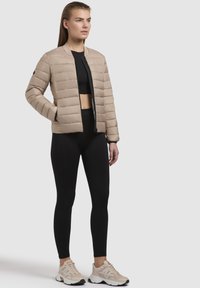 Giacca imbottita beige con un design corto, zip frontale, colletto a coste e maniche lunghe, abbinata a leggings neri e sneakers chiare.