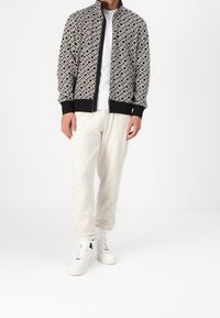 Chaqueta bomber a rayas en blanco y negro con cierre frontal, combinada con pantalones ligeros beige con un diseño de diamantes y zapatillas blancas.