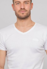 T-shirt en coton blanc avec un décolleté en V, des manches courtes, une coupe ajustée et une marque discrète d'une teinte légèrement plus claire sur la poitrine.