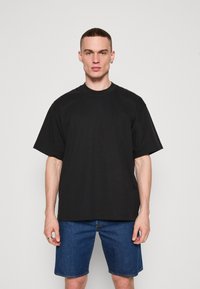 Tricou din bumbac negru cu guler rotund și mâneci scurte, având un detaliu subtil al logo-ului pe piept, asociat cu pantaloni scurți din denim.