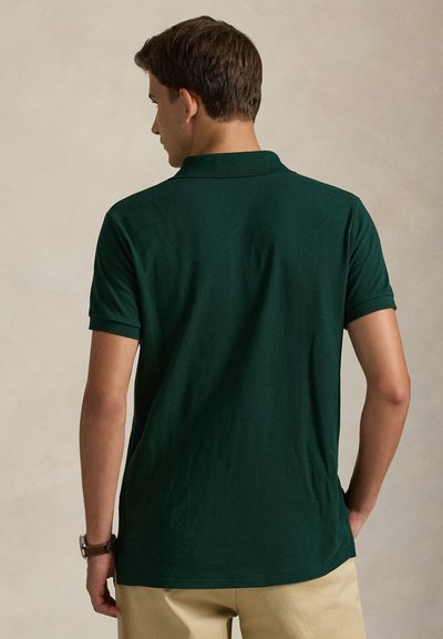 Mand iført mørkegrøn kortærmet poloshirt og beige bukser, vist bagfra mod en ensartet lys baggrund.