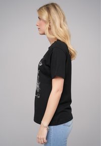 T-shirt en coton noir à manches courtes et à encolure ronde, avec un design graphique blanc sur le devant. Porté avec un jean bleu clair.