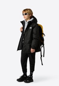 Black waterproof jacket with hood, gray sweatshirt, black pants, and black shoes. Yellow backpack, gray sunglasses, and logo accents visible.