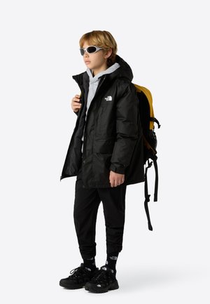 Schwarze wasserdichte Jacke mit Kapuze, grauer Sweatshirt, schwarze Hose und schwarze Schuhe. Gelber Rucksack, graue Sonnenbrille und sichtbare Logo-Details.