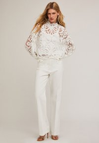 Witte kant blouse met luchtige mouwen en een hoge halslijn, gecombineerd met strakke witte broeken met knoopdetails. Hak sandalen maken de look af.