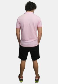 Polo shirt rosa con colletto e rifiniture delle maniche a contrasto, abbinato a pantaloni corti neri. Il modello sta in piedi con le mani in tasca, mostrando un look casual.