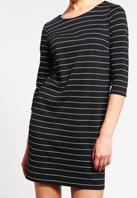 Svart klänning med vita horisontella ränder, trekvartsärmar, rund halsringning och avslappnad passform. Slät tyg med elastisk textur.