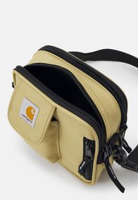 Beżowa torba na ramię z tkaniny z podwójnymi suwakami, czarnymi akcentami, przednią kieszenią i naszywką z logo Carhartt. Regulowany pasek do noszenia.