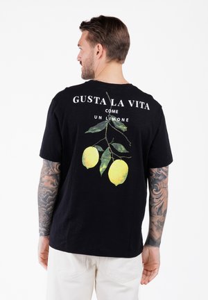 Hombre con brazos tatuados que lleva una camiseta negra con dos limones amarillos y texto italiano en la parte posterior, combinada con pantalones blancos.