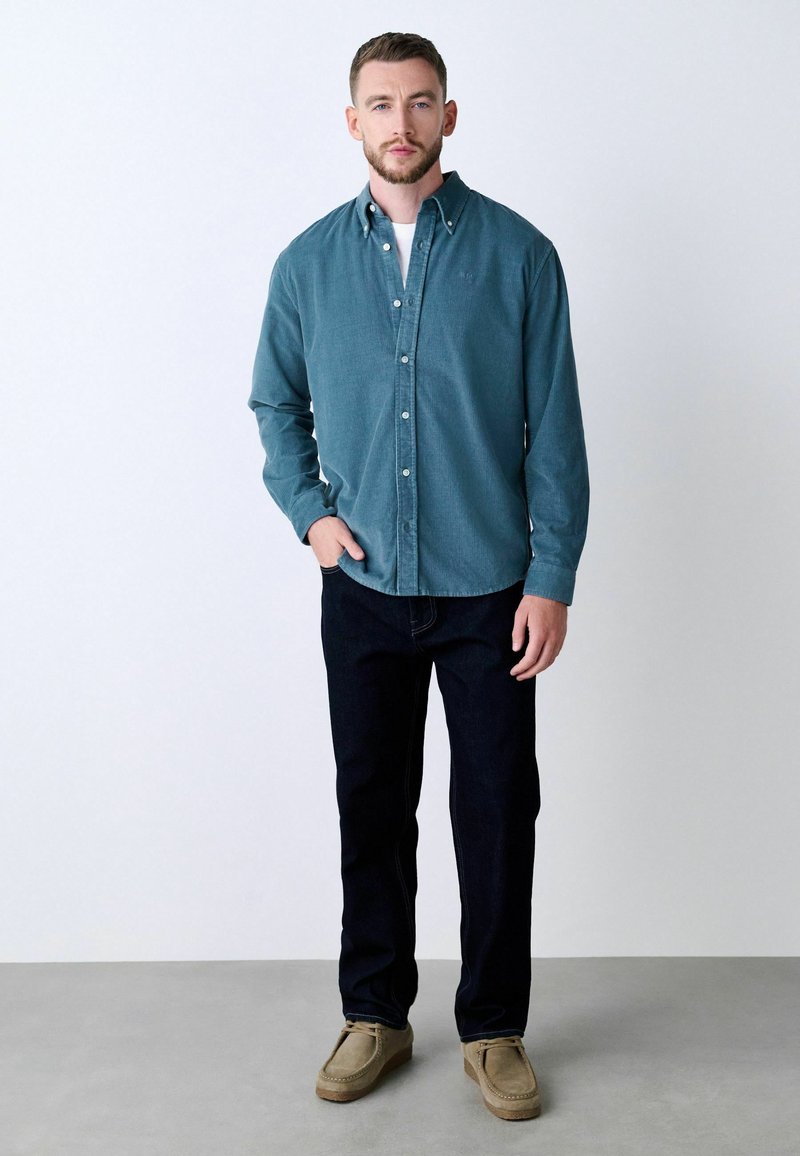 Cămașă cu nasturi din țesătură teal, cu mâneci lungi și guler, asortată cu blugi negri și pantofi bej. Design simplu, fără modele vizibile.