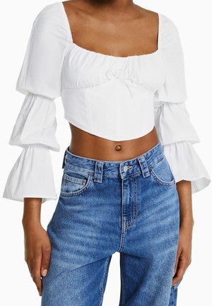 Kobieta w białej przyciętej bluzce z warstwowymi bufkami na rękawach i niebieskich jeansach z wysokim stanem, stojąca z rozluźnionymi ramionami.