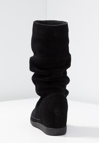 Botte montante en daim noir avec un design doux et froncé, bout rond et talon compensé. Texture lisse, détails minimalistes et couleur unie.