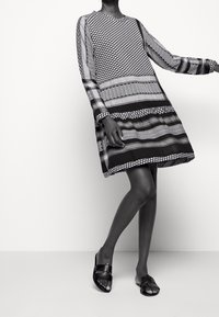 Schwarz-weiß gemustertes Kleid mit geometrischen Drucken, langen Ärmeln, rundem Ausschnitt und lockerem Schnitt. Kombiniert mit flachen schwarzen Sandalen.