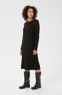 Femme aux cheveux bouclés portant une longue robe en tricot noire et des bottes en cuir noires jusqu'aux mollets avec des boucles, se tenant contre un fond uni.