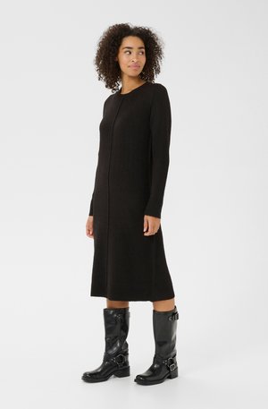 Femme aux cheveux bouclés portant une longue robe en tricot noire et des bottes en cuir noires jusqu'aux mollets avec des boucles, se tenant contre un fond uni.