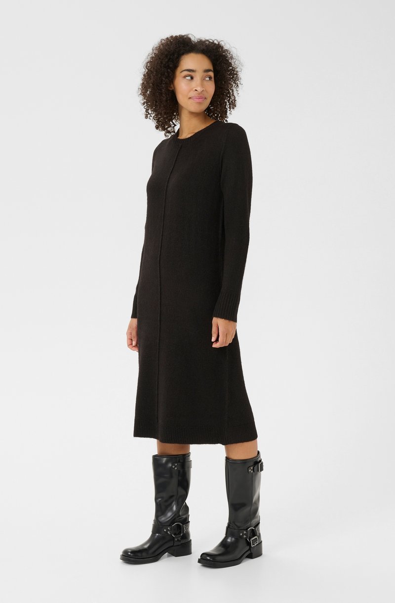 Femme aux cheveux bouclés portant une longue robe en tricot noire et des bottes en cuir noires jusqu'aux mollets avec des boucles, se tenant contre un fond uni.
