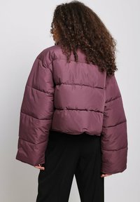 Chaqueta corta acolchada en burdeos profundo, con mangas amplias, textura acolchada y dobladillo redondeado. El material parece brillante y aislante.