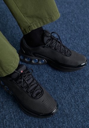 Person trägt schwarze Sportsneaker mit sichtbarer Luftdämpfung auf blauem Teppich, mit olivgrüner Hose und schwarzen Socken.
