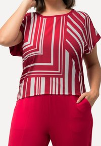 Rød T-shirt med korte ærmer, der har et geometrisk mønster med hvide striber. Glat stof med en afslappet pasform, som er parret med ensfarvede røde bukser.
