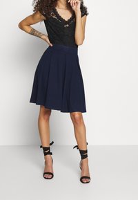 Frau mit einem schwarzen Spitzentop, einem marineblauen Faltenrock, schwarzen Ankle-Tie-Absätzen und einem gemusterten Tattoo am linken Unterarm, steht vor einem weißen Hintergrund.