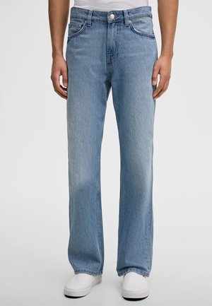 Osoba ubrana w jasnoniebieskie, proste dżinsy i białe wsuwane buty, stojąca na jednolitym tle.