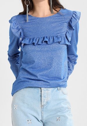 Kvinde iført en glitrende blå bluse med lange ærmer og flæser over bryst og skuldre, kombineret med lyseblå jeans med små broderede detaljer.