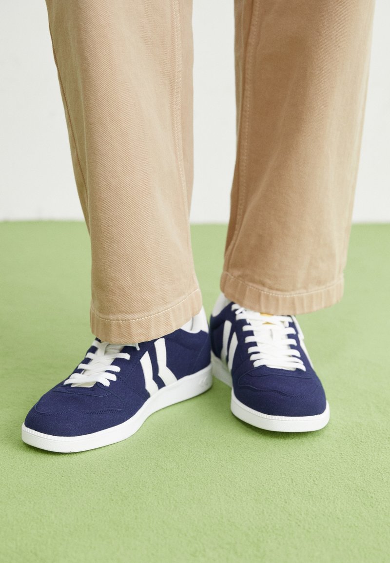 Personne portant un pantalon beige et des baskets bleu marine avec des lacets blancs et des rayures, debout sur un sol vert clair.