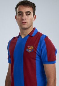 Rood en blauw gestreepte polo met een V-hals en korte mouwen. Bevat een geborduurd FC Barcelona-embleem op de linkerkant van de borst.