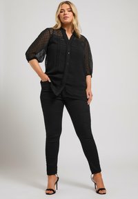 Lager svart blus med genomskinliga ärmar med prickar, knäppt front och plisserad detalj i kombination med slim-fit svarta byxor och remsandaler.