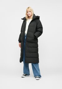 Hosszú fekete steppelt kabát kapucnival, amely steppelt mintázattal és oldalsó zsebekkel rendelkezik, világoskék bőszárú farmernadrággal és fekete cipővel párosítva.