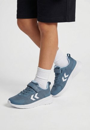 Benen met zwarte shorts, witte sokken en blauwe sneakers met witte chevron-logo's en klittenbandsluitingen op een witte achtergrond.