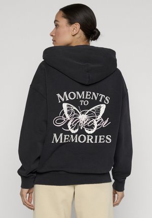 Femme aux cheveux en chignon portant un sweat à capuche noir avec un papillon blanc et le texte « Moments to Memories » au dos, pantalon beige visible.