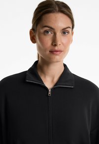 Schwarze zip-up Jacke mit gerippter Textur, hohem Kragen und einem silbernen Reißverschlussakzent. Schlichtes, modernes Design ohne zusätzliche Muster.