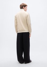 Μπεζ ζακέτα με ψηλό γιακά και απαλό ύφασμα, με εφαρμοστό λάστιχο στη μέση. Συνδυάζεται με χαλαρά μαύρα παντελόνια και ελαφριά παπούτσια.