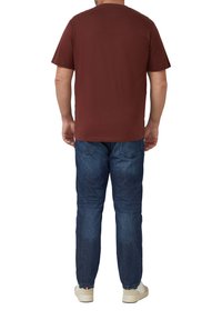 Brązowy krótki rękaw t-shirt, materiał bawełniany, okrągły dekolt, luźny fason. Widziany od tyłu, noszony z niebieskimi dżinsami i jasnymi trampkami.