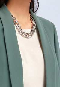 Collier chaîne en argent avec des maillons entrelacés, porté sur un haut clair, associé à un blazer vert texturé. Finition métallique lisse.
