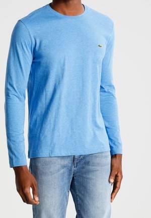 Langærmet blå bomulds T-shirt med rund halsudskæring og et lille, broderet logo på venstre bryst. Båret med lyse jeans.