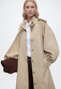 Γυναίκα που φοράει μια μπεζ καμπαρντίνα πάνω από λευκό πουκάμισο και σκούρο παντελόνι, κρατώντας ένα διπλωμένο σκούρο καφέ υφασμάτινο αντικείμενο στο ένα χέρι.