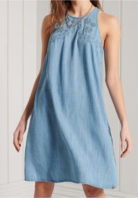 Vestido de mezclilla azul claro con un ajuste relajado, presenta bordados florales en el escote y un dobladillo redondeado. Diseño sin mangas.