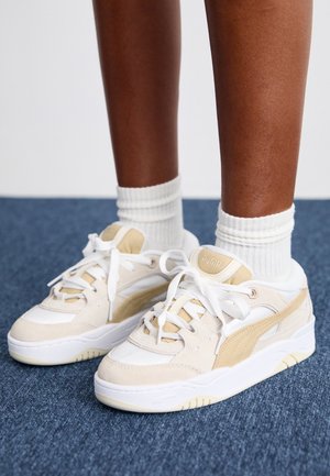 Person wearing white and beige Puma sneakers with white socks standing on a blue carpeted floor.