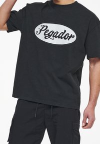 Černé oversized tričko vyrobené z bavlny, s velkým bílým oválem a nápisem "Pegador" v kurzívě. Jednoduchý design, krátké rukávy, kulatý výstřih.