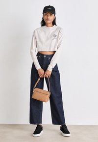 Haut blanc court à manches longues avec logo noir, pantalon large taille haute en denim foncé, baskets noires et petit sac à main beige.