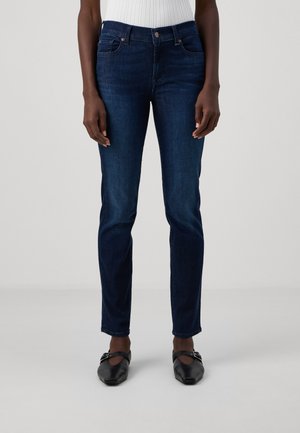 Temno modri oprijeti kavbojke z gladko denim teksturo, zasnovo s petimi žepi in srednje visokim pasom. Nosi se z črnimi ravnimi čevlji.