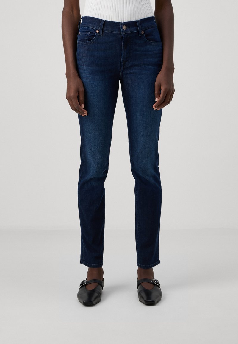 Donkerblauwe skinny jeans met een gladden denimtextuur, vijf zakken en een normale taille. Draagt met zwarte platte schoenen.