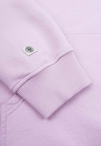 Ljust lila sweatshirtärm med ribbad textur och en liten tygetikett med en logotyp. Slät tyg, enkla design detaljer.