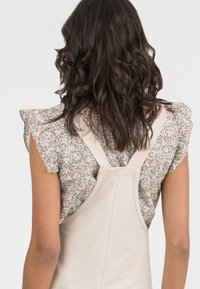 Beige overall dress with adjustable straps, paired with a floral-patterned blouse featuring ruffled sleeves. Long, wavy dark hair visible.