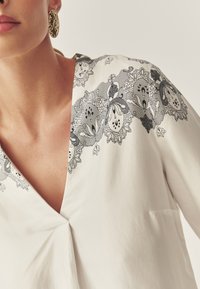 Blouse blanche à col en V profond, ornée de broderies florales noires le long de l'encolure. Le tissu semble lisse et léger.