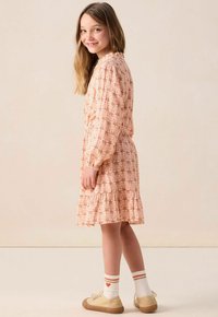 Rosa gemustertes Kleid mit Rüsche am Saum, langen Ärmeln und hohem Kragen. Kombiniert mit weiß-rot gestreiften Socken und goldenen Sneakers.